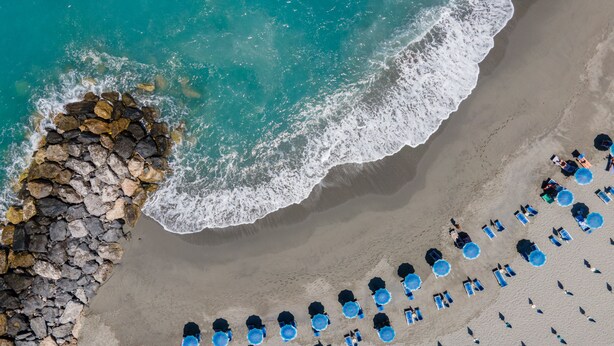 Luchtbeeld van een strand met een rij blauwe parasols en ligbedden, een steiger en de golven van de oceaan.