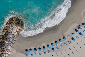 Luchtbeeld van een strand met een rij blauwe parasols en ligbedden, een steiger en de golven van de oceaan.