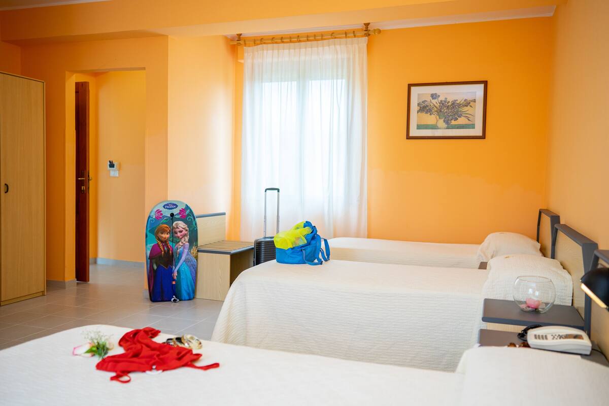 Een hotelkamer met twee bedden, een raam met dunne gordijnen, een nachtkastje met een lamp, en een lijst met een afbeelding aan de muur.