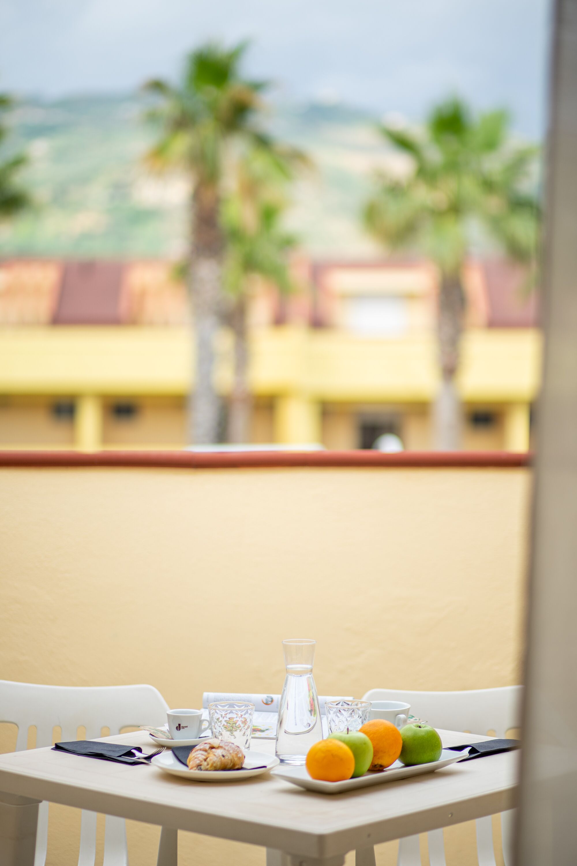 A table with a plate of fruit, a glass of water, and a pastry, set against a yellow wall with a view of palm trees and buildings.