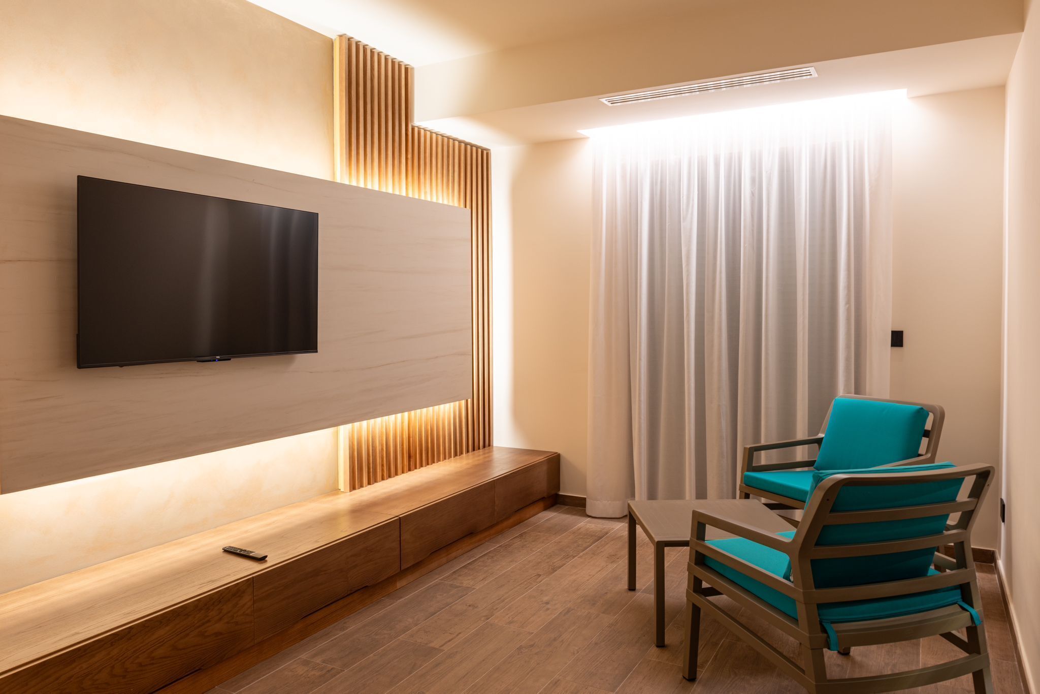 A modern room with a flat-screen TV mounted on a wooden wall, a small table with chairs, and a large window with curtains.