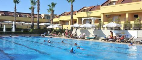 Una piscina con múltiples carriles, rodeada de sillas de descanso y sombrillas, con un edificio con balcones y palmeras al fondo.