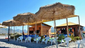 Een restaurant aan het strand, met rieten parasols, witte plastic stoelen en een houten gebouw.
