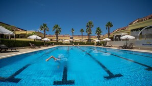 Een zwembad met helderblauw water, omringd door ligbedden en parasols, met palmbomen en een hotelgebouw op de achtergrond.