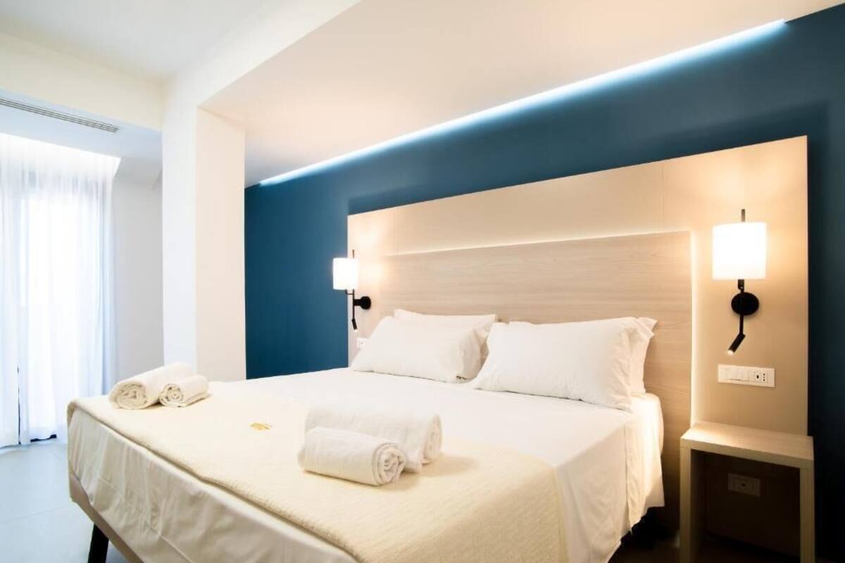 Een opgemaakt bed met witte lakens en opgerolde handdoeken, een nachtkastje met een lamp, en een wandlamp.
