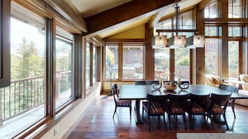 A spacious dining area with a large table and chairs, large windows offering views of the outdoors, and a wooden ceiling.