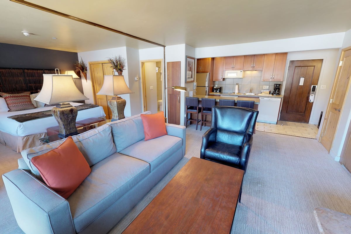 Una sala moderna con un sofá, sillones y una mesa de centro. Cuenta con una zona de cocina con armarios de madera y un microondas.