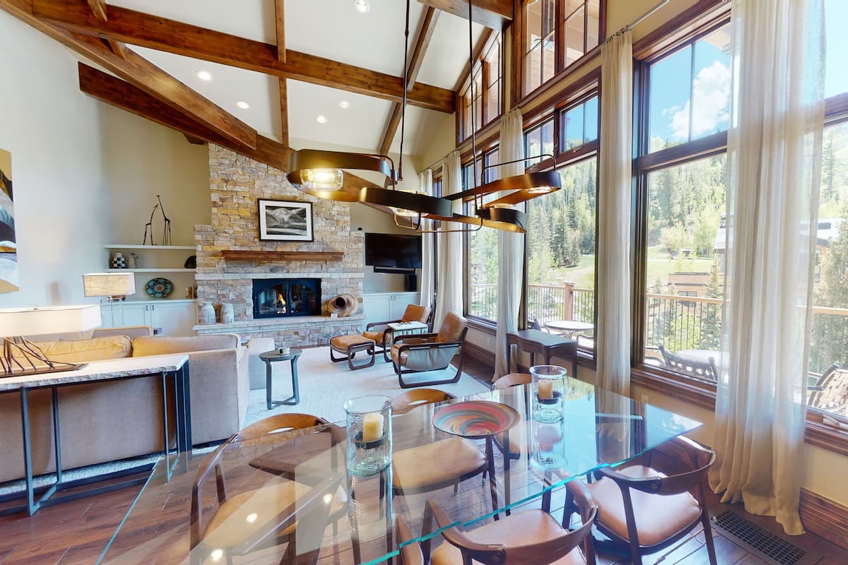 Amplia sala de estar con chimenea de piedra, vigas de madera y ventanas grandes que ofrecen vistas al exterior.