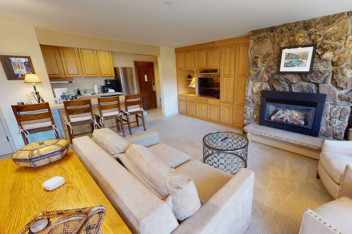 Una sala de estar con chimenea de piedra, sofás color beige y una zona de cocina con armarios de madera y un refrigerador.