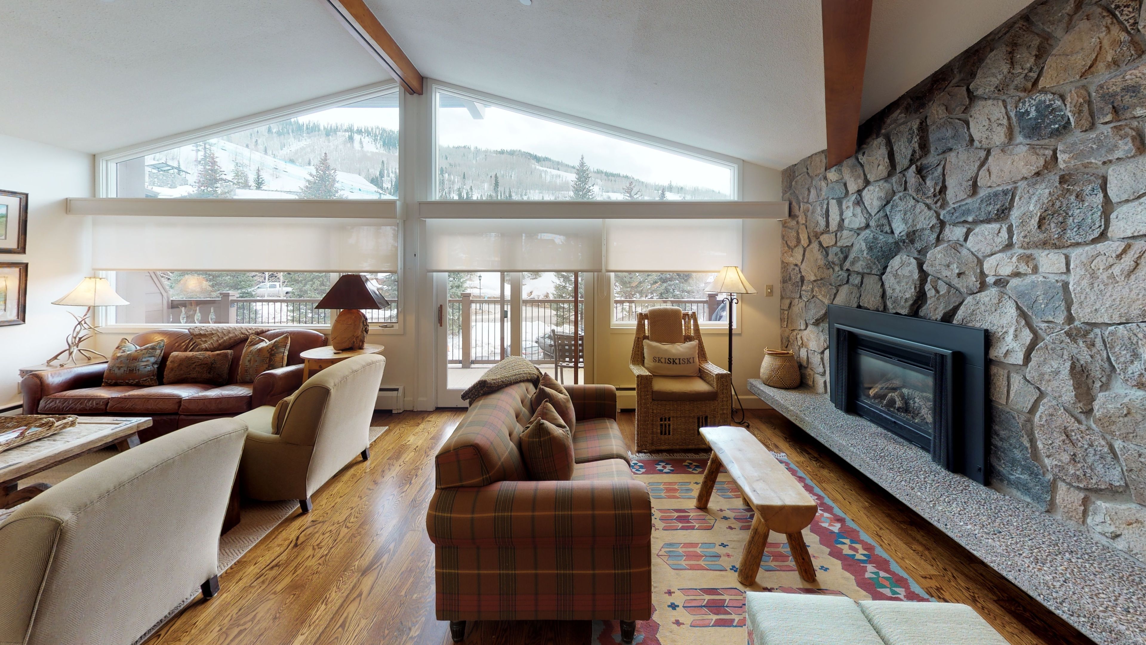 Un salon chaleureux avec une cheminée en pierre, de grandes fenêtres et une vue sur des montagnes enneigées.