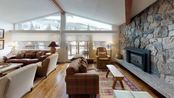A cozy living room with a stone fireplace, large windows, and a view of snow-covered mountains.
