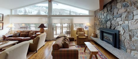 A cozy living room with a stone fireplace, large windows, and a view of snow-covered mountains.