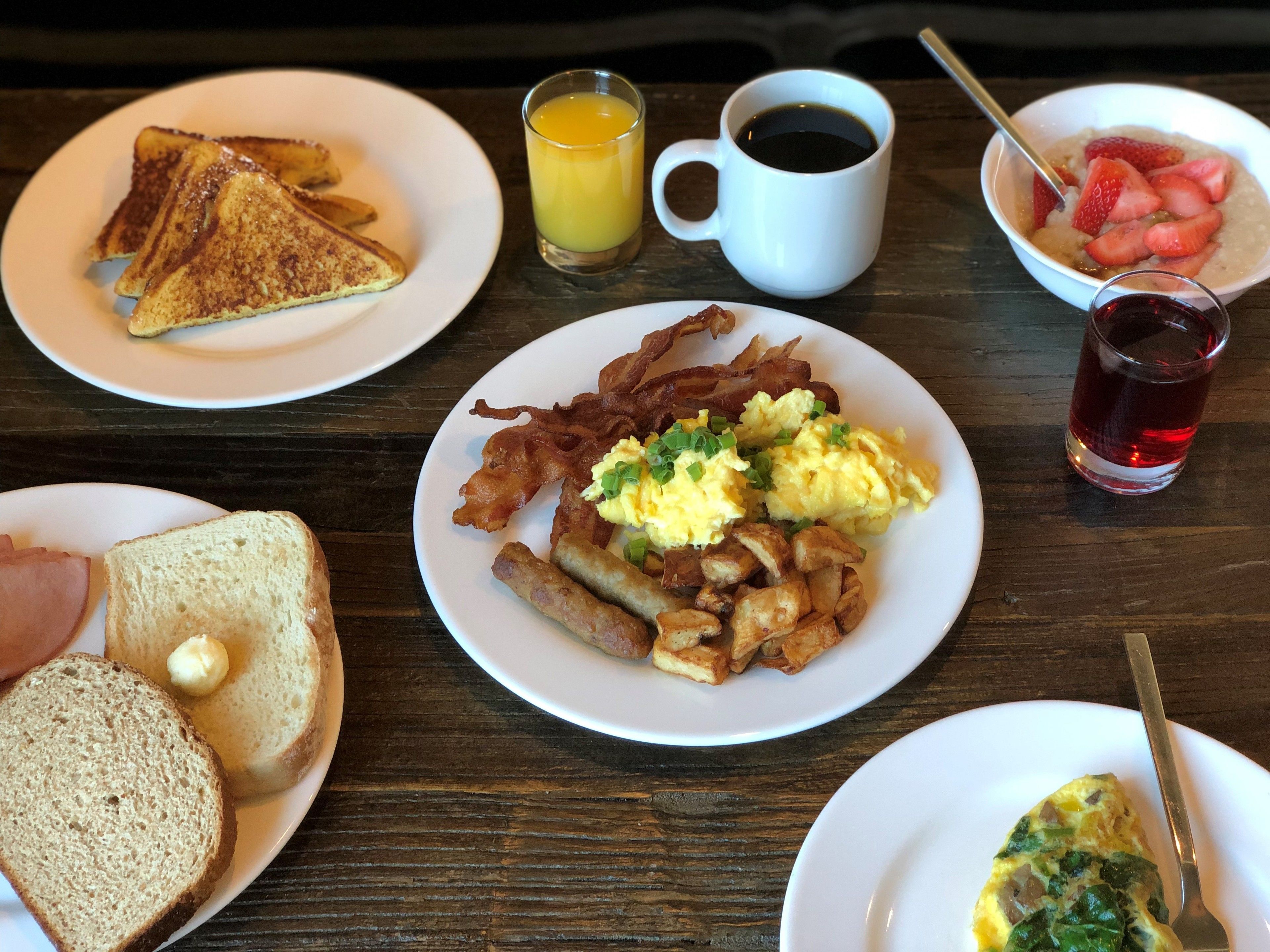 A breakfast table with toast, scrambled eggs, bacon, hash browns, a cup of coffee, a glass of orange juice, a bowl of strawberries, and a glass of red beverage.