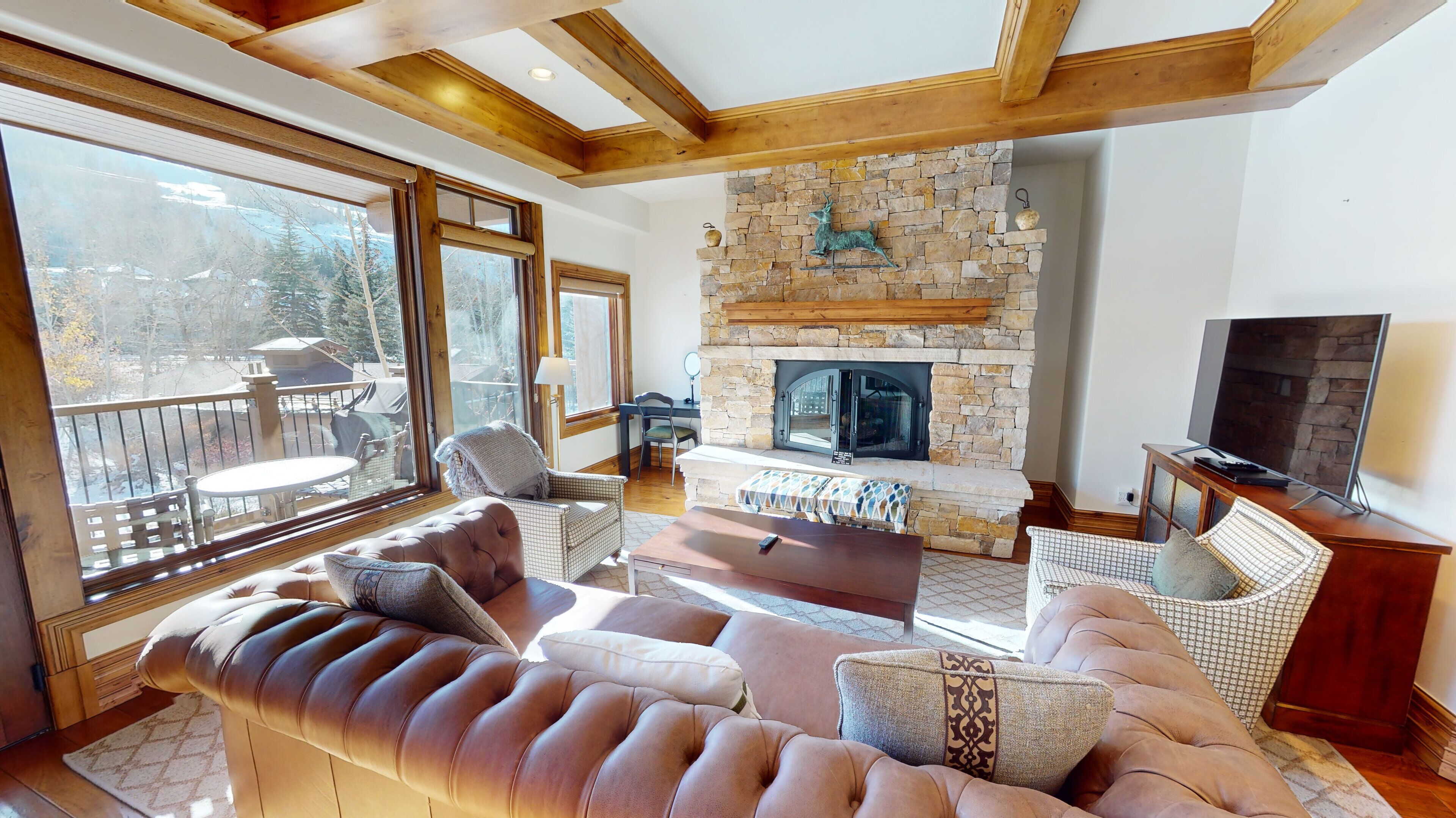 A cozy living room with a stone fireplace, comfortable seating, and large windows offering a view of the outdoors.