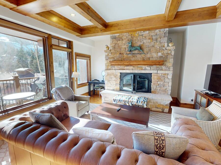 A cozy living room with a stone fireplace, comfortable seating, and large windows offering a view of the outdoors.