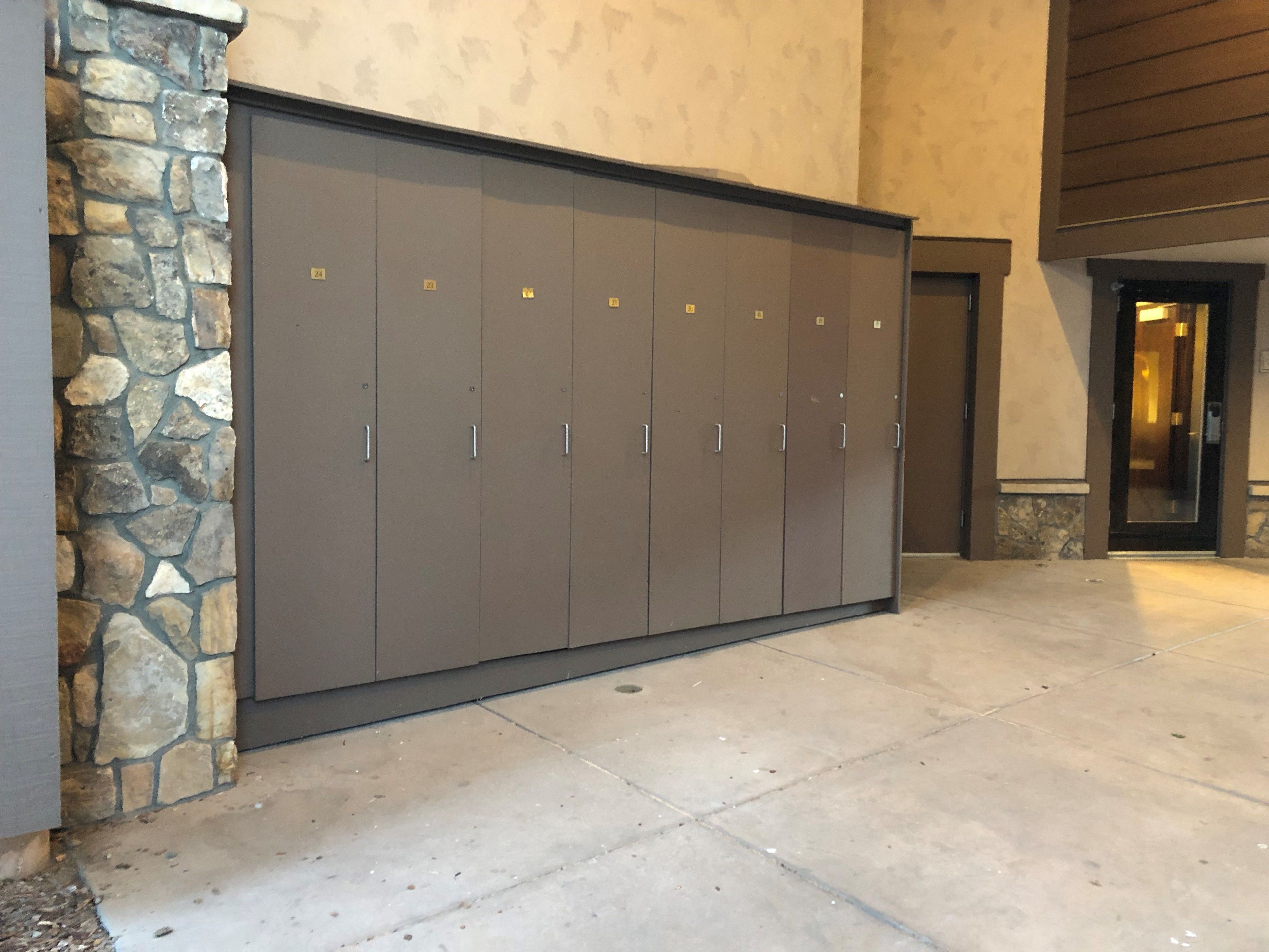 Een rij grijze lockers met metalen handgrepen, geplaatst tegen een stenen muur en een inkomhal.