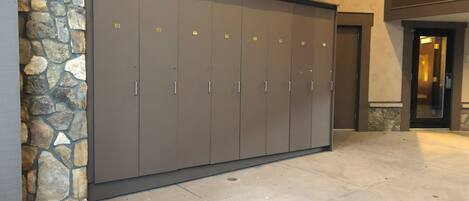 A row of grey lockers with metal handles, set against a stone wall and a building entrance.