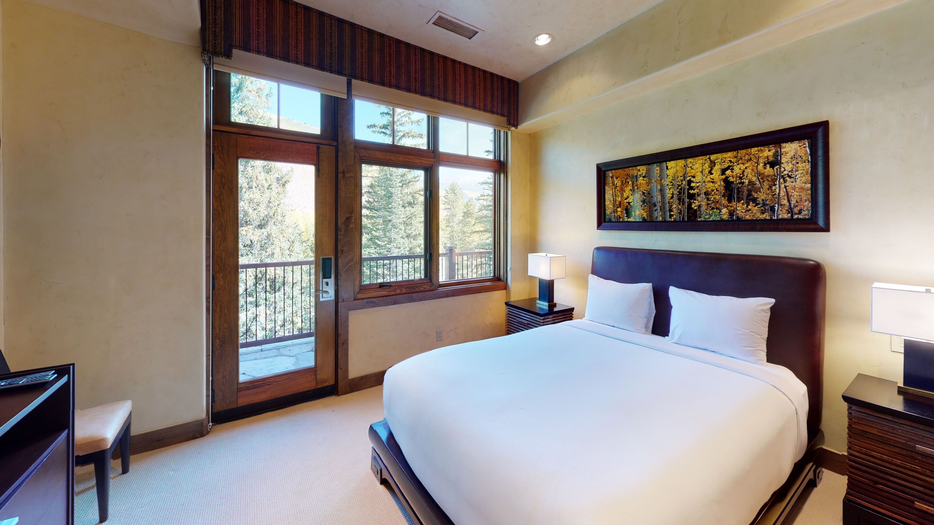 A hotel room with a large bed, a desk, a chair, a nightstand with a lamp, and a painting on the wall.