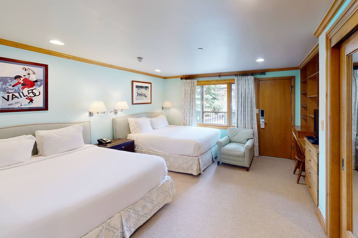 Habitación de hotel con dos camas, un sillón pequeño, un escritorio de madera, un televisor y una ventana con cortinas.
