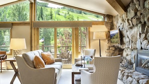 A cozy living room with a stone fireplace, large windows offering a view of a green landscape, and comfortable seating.