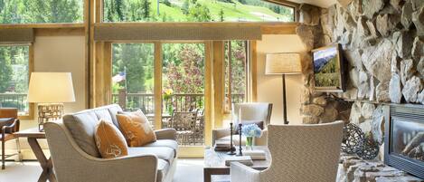 A cozy living room with a stone fireplace, large windows offering a view of a green landscape, and comfortable seating.