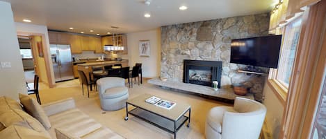 A modern living room with a stone fireplace, a flat-screen TV mounted above it, and a dining area with a kitchen in the background.
