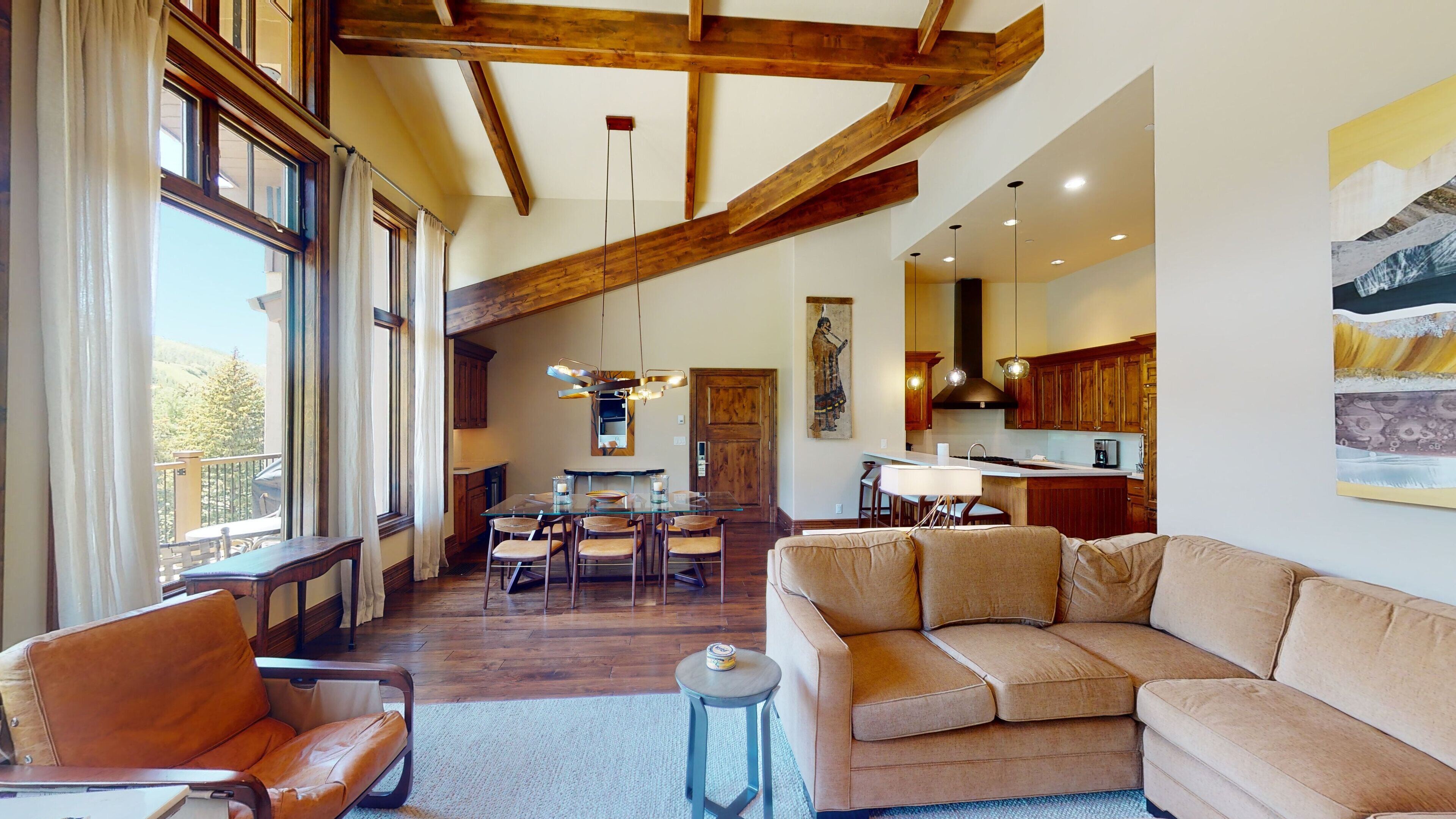 A spacious living area with a large window, wooden beams, a dining table, and a sofa.