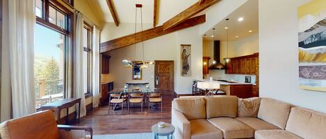 A spacious living area with a large window, wooden beams, a dining table, and a sofa.