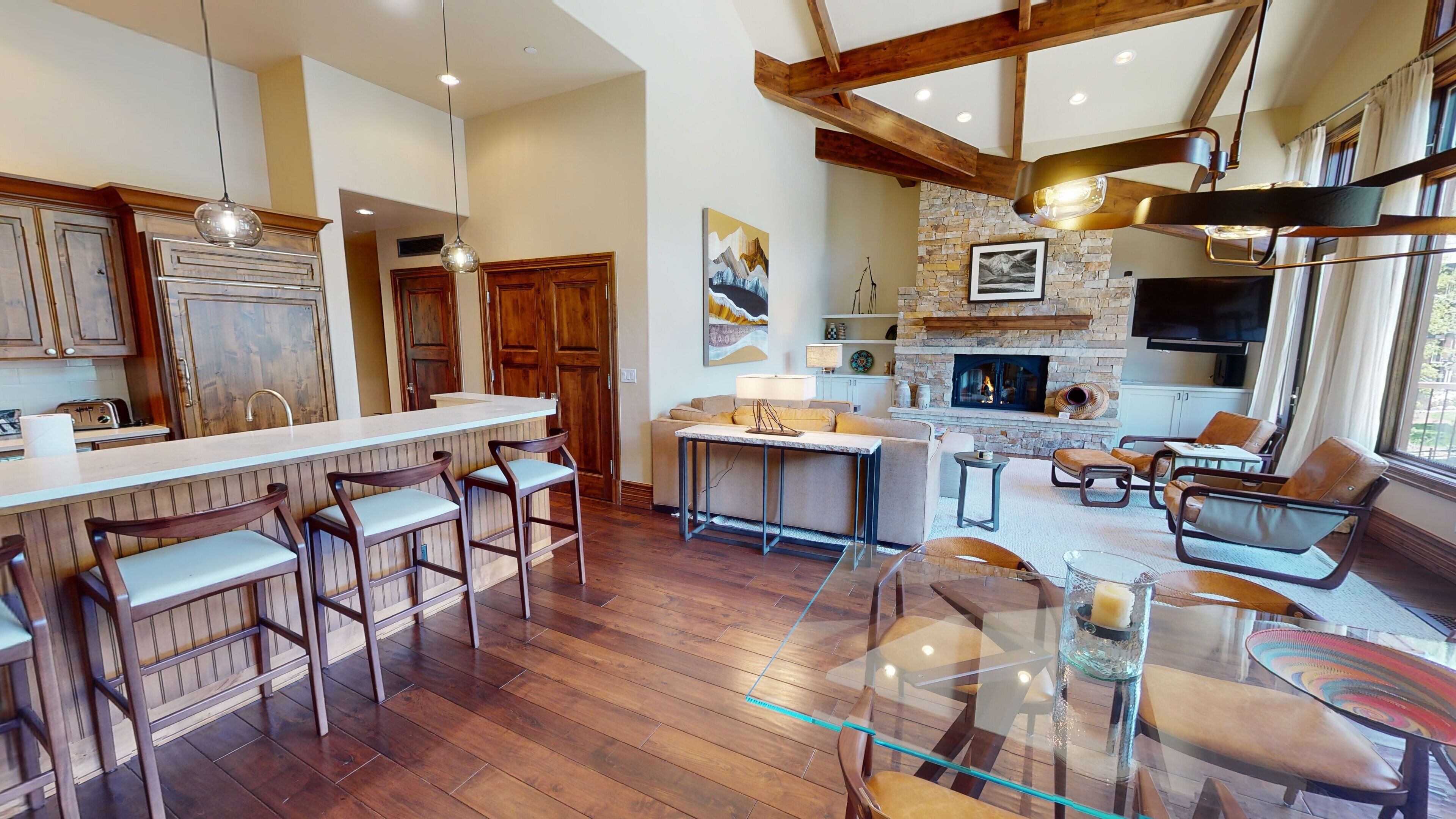 Une cuisine spacieuse avec des meubles en bois, un coin bar avec des tabourets, une cheminée en pierre et un coin salon avec des canapés en cuir.