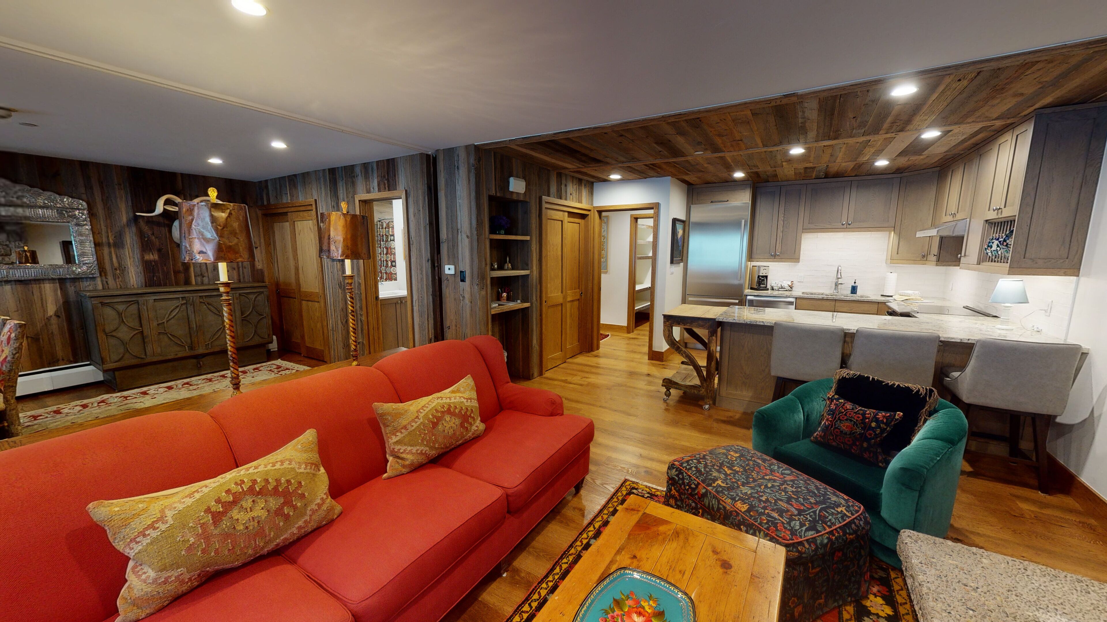 Una sala de estar moderna con un sofá rojo, un sillón verde azulado y una mesa de centro de madera. Se ve una cocina al fondo con armarios de madera y un refrigerador de acero inoxidable.