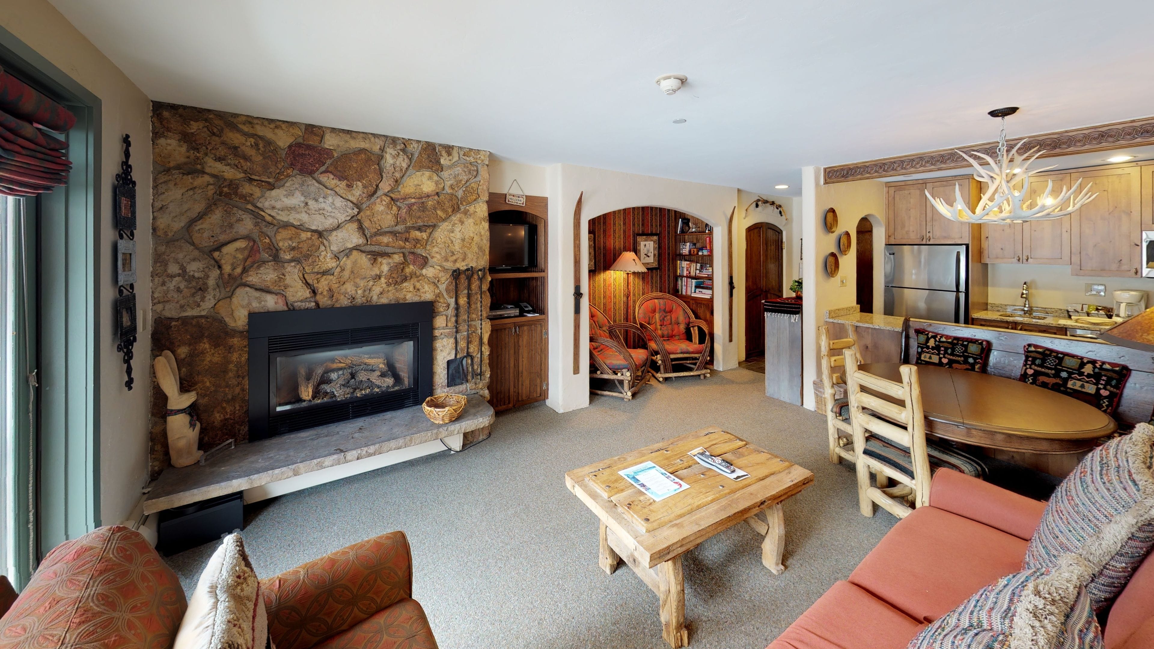 A cozy living room with a stone fireplace, a dining area with a wooden table, and a kitchen with a refrigerator.