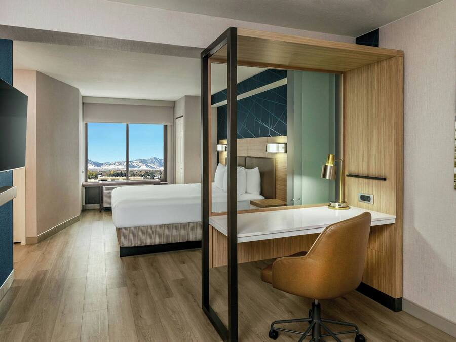 Habitación de hotel con una cama grande, un escritorio con silla, televisor y vistas a montañas a través de amplias ventanas.