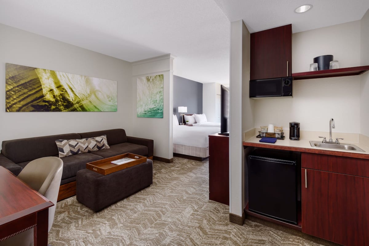 A hotel room with a sofa, ottoman, and a small dining table. There is a kitchenette with a microwave, sink, and cabinets. The room has a large window and artwork on the walls.