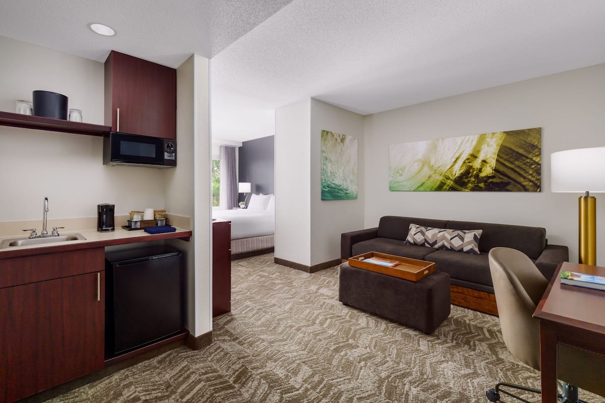 A compact hotel room with a kitchenette, a sofa, a desk, and a large abstract painting on the wall.
