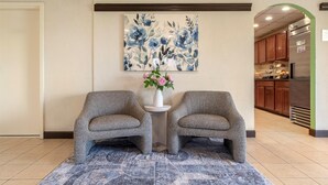 A lobby with two grey armchairs, a blue patterned rug, a floral vase, and a large abstract painting on the wall.