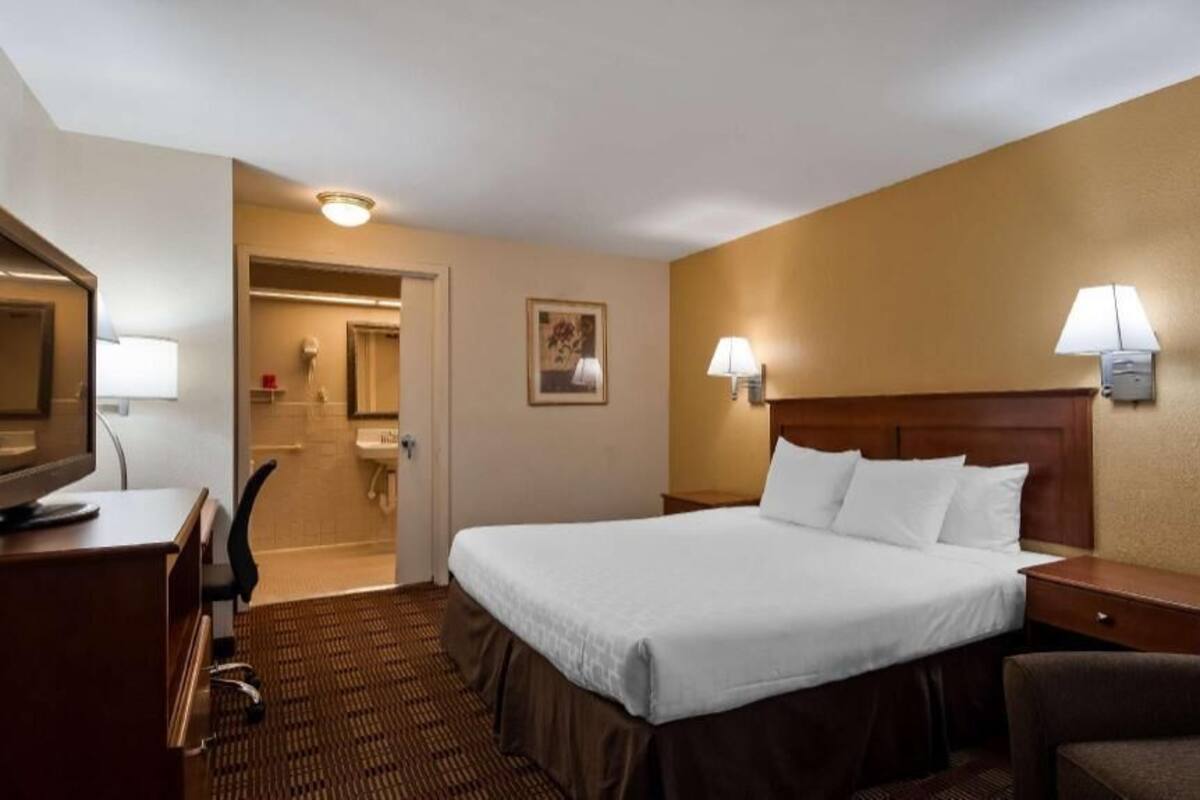 Ein Hotelzimmer mit Bett, Nachttisch, Lampe, Stuhl, Schreibtisch und einem durchgegangene Tür erkennbaren Badezimmer.