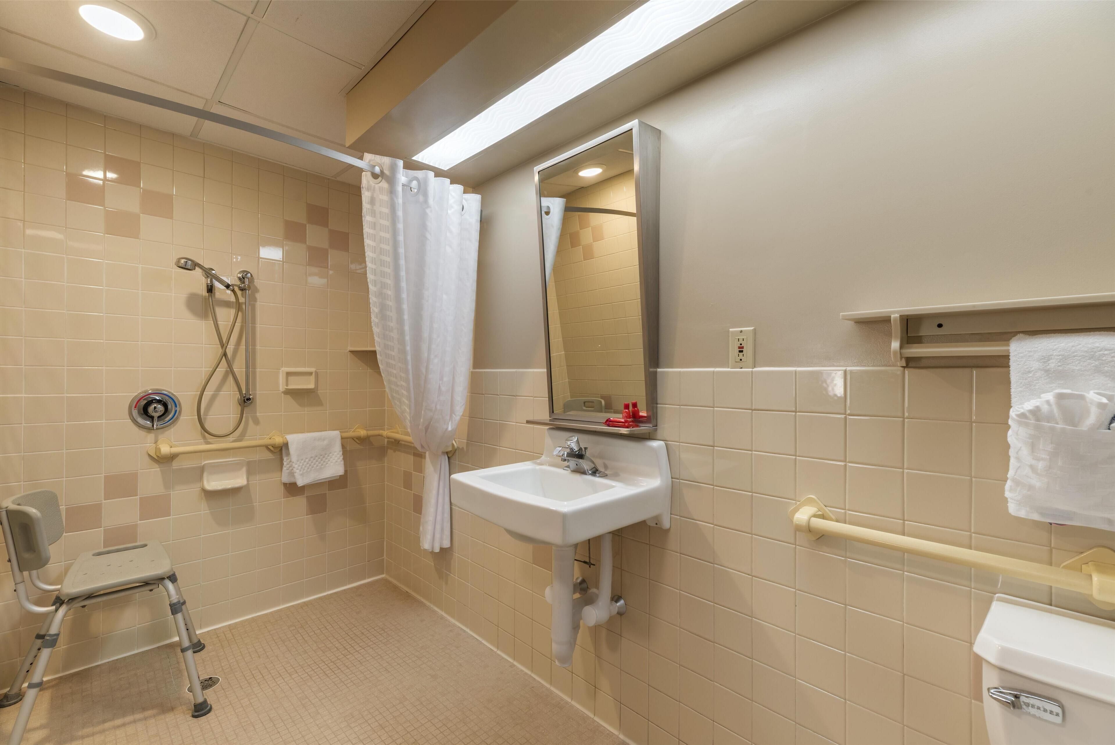 A bathroom with a shower, a sink, a mirror, and a wheelchair.