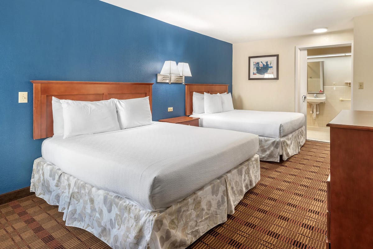 Habitación de hotel con dos camas, cabecero de madera, mesita de noche, lámpara, baño con lavamanos y espejo, y alfombra estampada.