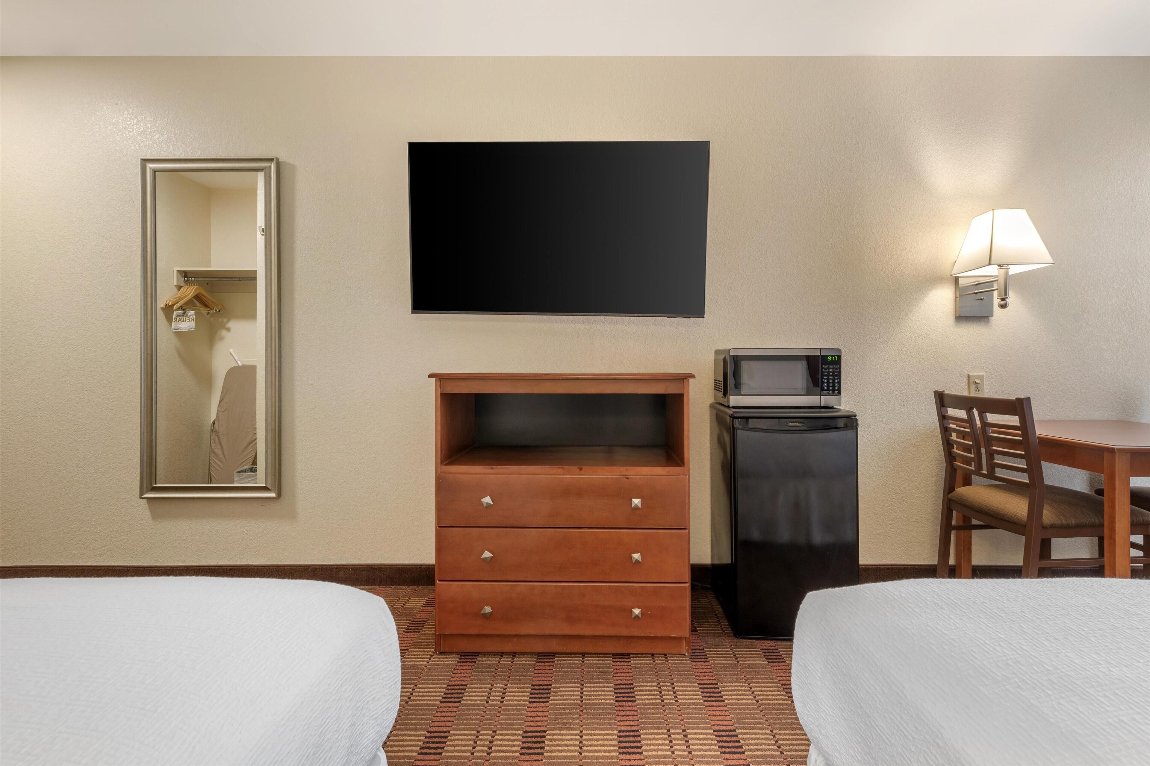 A hotel room with two beds, a wooden dresser, a flat-screen TV, a microwave, a desk with a chair, and a wall-mounted lamp.