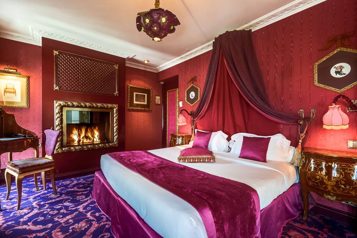 A luxurious bedroom with a large bed, ornate decor, a fireplace, and a chandelier.