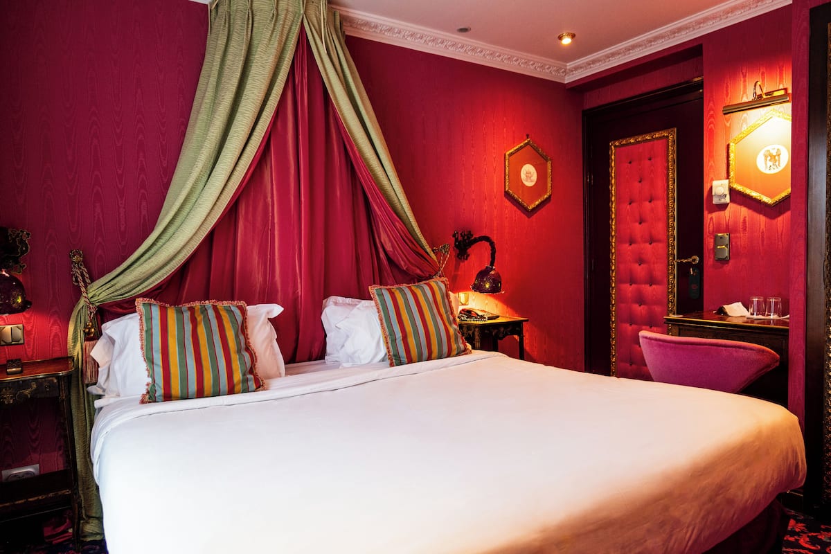 A large bed with white linens, a green canopy, and striped pillows. There is a nightstand with a lamp and a mirror on the wall.