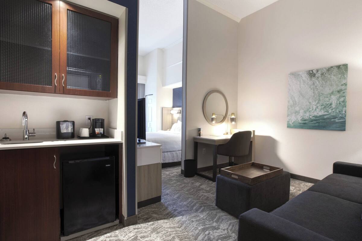 Habitación de hotel con kitchenette, una cama, un escritorio con silla, una mesita y un cuadro en la pared.