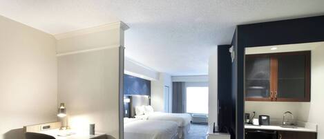 A hotel room with a desk, two beds, a kitchenette, and a large window.