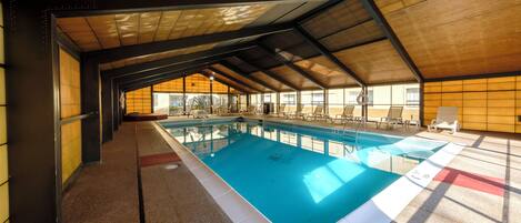 An indoor swimming pool with a tiled floor, surrounded by lounge chairs and a high, sloped ceiling.