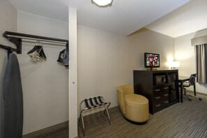 A hotel room with a bed, a TV, a desk, a chair, a small stool, and a coat rack with hanging items. - Red Roof Inn & Suites Newburgh – Stewart Airport/ West Point (New Windsor)