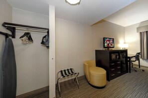 Habitación de hotel con cama, televisión, una mesita, una silla y perchero con prendas colgadas.
