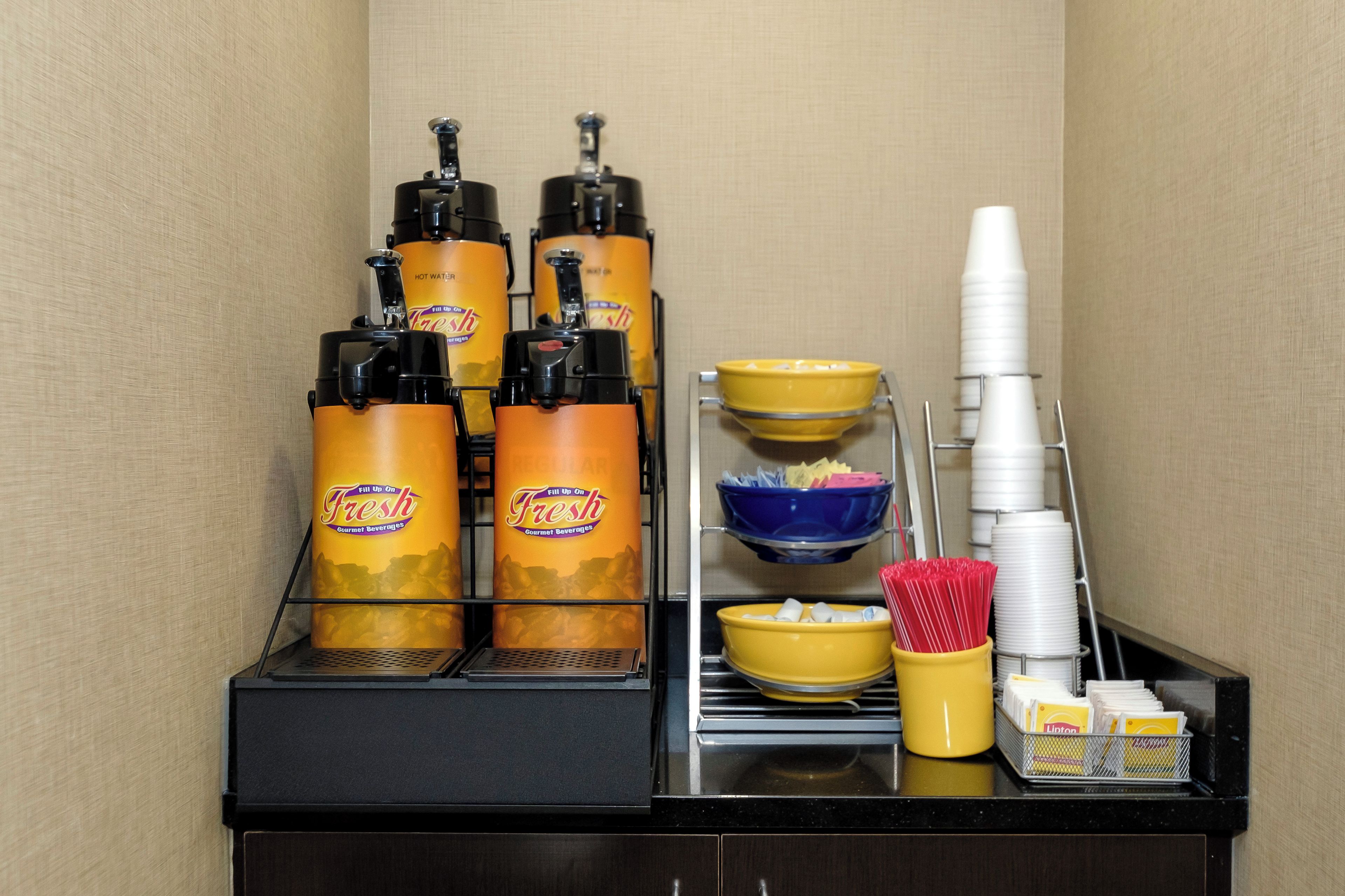 A beverage station with multiple dispensers, a stack of paper cups, and a tray with bowls and napkins.