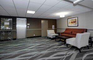 Una habitación de hotel con alfombra estampada, dos sillones, un sofá, un espejo grande y un cuadro abstracto enmarcado en la pared.