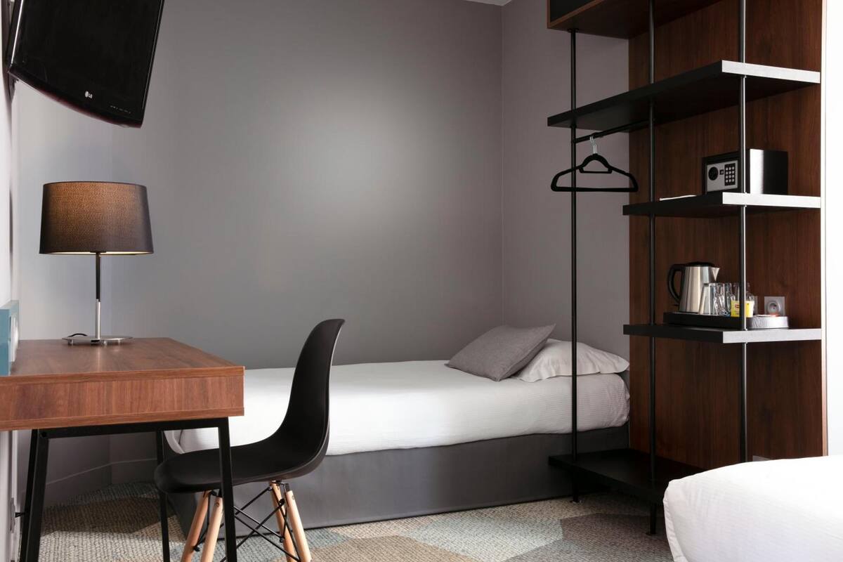 A compact hotel room with a bed, a desk with a chair, a TV mounted on the wall, and a shelf with a radio and a kettle.