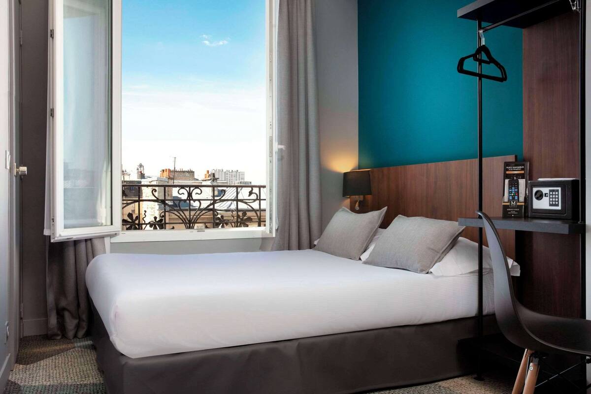 A hotel room with a bed, a window with a view of buildings, a hanging coat rack, and a bedside table with a lamp and a phone.